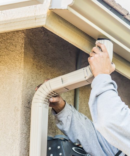 Worker Attaching Aluminum Rain Gutter and Down Spout to Fascia of House